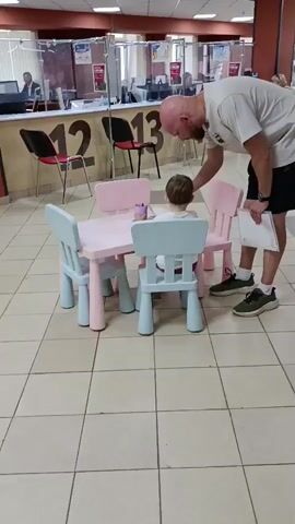A bald man in a white t-shirt and black shorts is bending over to interact with a young child sitting at a small pink and blue table. The child is seated in a blue chair, facing away from the camera, and appears to be holding a purple cup and possibly drawing or playing with something on the table. The man is holding a white document in his left hand and is gently touching the child's head with his right hand. The scene appears to be set in a public waiting area, possibly a government office or a service center, with numbered booths and partitions visible in the background. Other people can be seen in the background, some waiting in line and others behind counters. The floor is tiled.