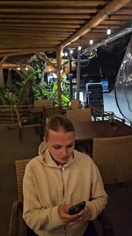 A woman in a light brown hooded sweatshirt is sitting at a table. She is holding a black smartphone and looking at it. The woman then puckers her lips and makes a kissy face towards the camera. The scene appears to be outdoors at night. There are wooden tables and chairs, with a thatched roof overhead. Lights are strung across the roof, and there is lush green foliage in the background.