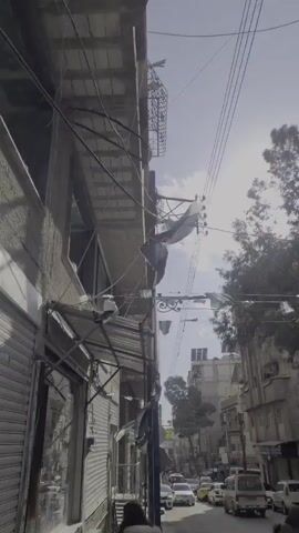The video shows a street in what appears to be a Middle Eastern city, with the Syrian flag prominently displayed. The flag is hanging from a power pole attached to a building.  Smaller Syrian flags are strung across the street, creating a festive or celebratory atmosphere.  The buildings are older and show signs of wear.  Power lines are visible, creating a cluttered appearance. The sky is overcast with hints of sunlight breaking through.  Overall, the scene suggests a location where national identity is being asserted or celebrated amidst a backdrop of urban decay.