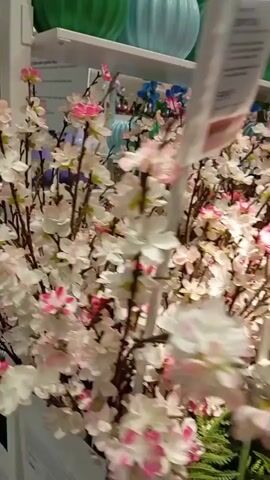 The video shows a close-up view of artificial flowers displayed in what appears to be a store or market stall. The flowers are arranged in bunches and include various types and colors, such as white and pink cherry blossoms, green ferns, pink orchids, blue hydrangeas, and yellow sunflowers. Some of the flowers have price tags or informational labels attached to them. The camera slowly pans across the display, giving a detailed look at the artificial flora. The lighting is bright, suggesting an indoor environment.