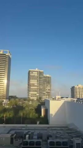 The video shows a cityscape on a clear, sunny day. In the foreground, there's a white, flat rooftop covered with air conditioning units. Beyond that, there is a parking structure with many cars parked on it. In the background, there are several tall, modern buildings. Some greenery, possibly palm trees, is visible at the base of the buildings. The sky is blue with a few scattered clouds. The camera slowly pans, revealing more of the parking structure and the buildings in the background.