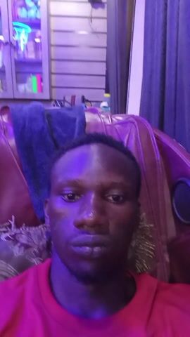 A close-up video shows a dark-skinned man in a red T-shirt. He is sitting in a red leather chair in a room with violet-blue lighting. Behind the chair is a cabinet displaying various items. The man looks directly at the camera throughout the video.