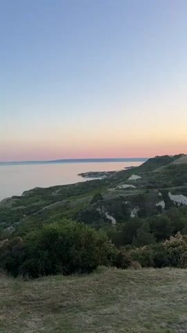 This is a vertical, time-lapse video showing a sunset over a coastal landscape. The scene begins with a view of rolling green hills and a body of water under a clear, pale blue sky. As the video progresses, the sky transitions through shades of pink and orange as the sun dips below the horizon. The landscape is covered in dense green vegetation, with some areas of lighter-colored soil visible on the hillsides. In the foreground, there is dry grass and a few small bushes. The video captures the changing light and colors of the sunset, with the sun appearing as a bright orange disc near the end of the clip.