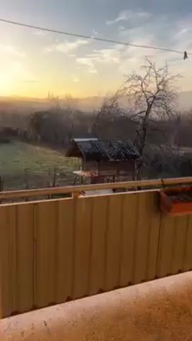 The video shows a sunrise over a rural landscape. The sun, a brilliant yellow-white orb, is visible on the horizon, partially obscured by the silhouette of hills and trees. The light from the sun illuminates the sky, creating a warm golden glow. In the foreground, there is a small, rustic wooden structure with a slanted roof. A yellow fence is also visible, running horizontally across the bottom of the frame, partially obstructing the view. The land beyond the fence is a green field, and further in the distance, outlines of hills fade into the bright, hazy background.