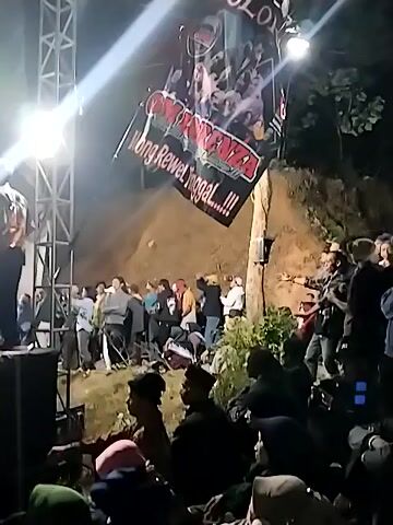 A video taken at night shows a crowd of people gathered outdoors. A large banner hangs diagonally from a tree, illuminated by bright lights. The banner has text and images on it. Several lights are also positioned on a metal truss structure in the foreground. People are visible in the background, some standing and some sitting or lying down, with a dirt incline behind them. The scene appears to be an event or performance.