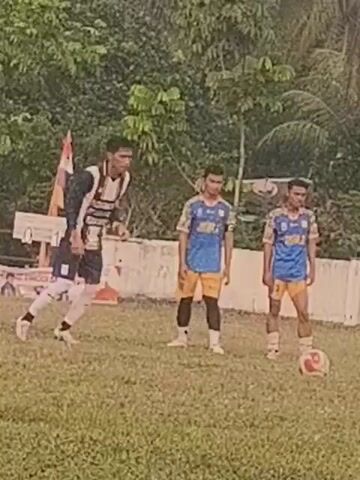 The video shows a group of young men playing soccer on a grassy field. There are two teams, one wearing blue and yellow uniforms, and the other wearing black and white uniforms. The men are kicking a soccer ball and running around the field. The scene appears to be outdoors, with trees and foliage in the background. The weather appears to be overcast, and there is a white fence in the background. One of the players in the black and white uniform appears to be the goalkeeper. The other players are playing offense and defense. The game appears to be in progress, with the players actively engaged in the sport. There are spectators in the background. One player in a black and white uniform appears to be the captain or leader. The players appear to be young adults or teenagers. The video is a close-up shot of the game, focusing on the players and the action.