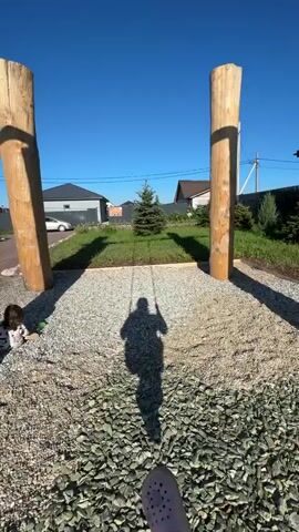 The video shows a first-person perspective of someone sitting on a swing. Two large wooden posts frame the view. In the background, there is a clear blue sky, houses, a small evergreen tree, and a white car parked in front of a house. The person is wearing pink Crocs and gray socks. The shadow of the person on the swing is visible on the gravel ground. A child is briefly visible in the lower left corner of the frame at 00:03. The swing is in motion, swinging forward and backward.