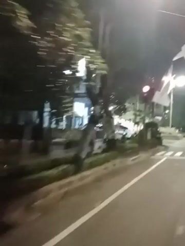 The video, captured at night, shows a street scene with a focus on a road and sidewalks. There are flags, possibly national flags, with red and white colors, displayed on poles, with one flag in particular being prominent and appearing to be a national flag. Several cars are visible on the road, some appearing to be parked and others in motion. Streetlights illuminate the scene, along with the headlights of the moving vehicles. A pedestrian crossing is marked on the road. Buildings and trees are present along the sides of the road. The camera appears to be moving, suggesting it is mounted on a vehicle. The overall impression is of a moderately busy street at night.