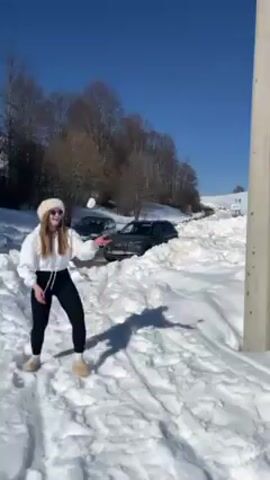 A young woman is standing in deep snow, wearing black leggings, a white sweatshirt, and a fuzzy white hat. She's also wearing light-colored, slip-on shoes or slippers. The scene is a snow-covered landscape on a sunny day with a clear blue sky. Behind her, there's a snow-filled roadway with a couple of cars partially buried or stopped in the snow. There are bare trees lining the side of the road, and a concrete utility pole is visible on the right edge of the frame. The woman makes gestures with her arms, seeming cheerful and playful in the snowy environment, then starts walking toward the snow-filled road.