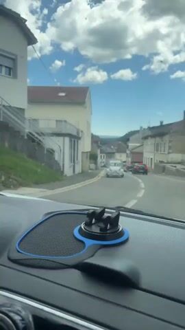 The video is shot from the perspective of someone in a car, looking out the windshield. The car is driving down a road in a town with buildings on either side. There are other cars on the road, including a vintage white car in the distance. The sky is blue with white clouds. The video is taken during the daytime.