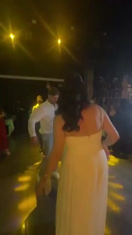 A man in a white shirt and light-colored pants dances on a dark floor. He is illuminated by blue and red stage lights. A woman in a white dress stands with her back to the camera, also illuminated by the lights. Other people are visible in the background. The scene appears to be a party or event.