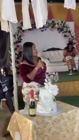 A woman speaking into a microphone stands next to a decorated table with a cake and bottle of champagne at a wedding reception. Behind her on a stage, a woman sits on a couch. A wedding arch of flowers is behind the couch. Curtains hang from the ceiling. A woman in a gown walks onto the stage.