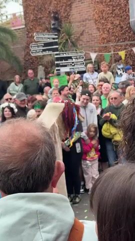 A crowd of people is gathered in an outdoor setting, possibly a town square or public space. The focus is on a figure draped in a large, rough-textured cloth, possibly burlap or a similar material, which obscures their body. The figure has a headpiece adorned with colorful ribbons or strips of fabric. People in the crowd are watching, smiling, and recording the event with their phones. A few individuals are wearing flower crowns, suggesting a festive or celebratory occasion. The environment includes brick buildings with climbing foliage, decorative flags, and directional signs indicating local landmarks. The atmosphere appears lively and cheerful, with the crowd's attention centered on the cloaked figure.