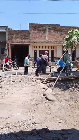 The video shows a group of men working on a construction site in front of a building. The building appears to be a house with a brick facade and a covered entryway. The construction site is rough and unpaved, with gravel and debris scattered on the ground. Several men are actively engaged in construction work, including what appears to be pouring concrete. One man is riding a motorcycle on the side of the road. The weather is clear and sunny.