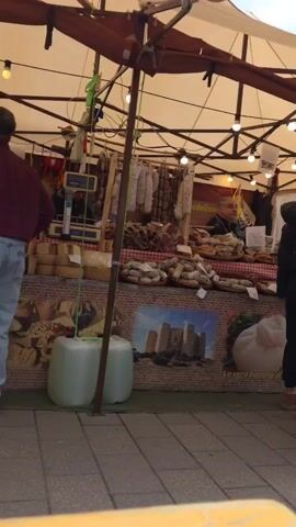 The video shows an outdoor marketplace in a town square. Several stalls are set up, selling Italian specialties like sausages and cheeses. The Italian flag is prominently displayed. People are browsing and walking around the stalls. A person sits on a bench near a stall selling Italian specialties. Another person pushes a child in a stroller. The architecture suggests a European setting, with a large church in the background. The weather appears to be overcast.