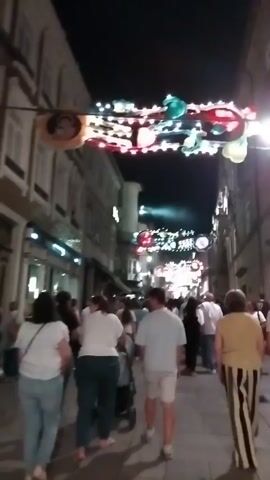 A nighttime street scene, viewed from behind a group of people walking away from the camera. The street is lined with buildings on both sides. Overhead, strings of festive lights and decorations are strung across the street. Several people are visible walking in the same direction as the camera, and others are seen in the distance. The lighting suggests it is dusk or evening. The people are dressed in casual attire. One person is pushing a stroller. The ground is paved with stones or cobblestones.