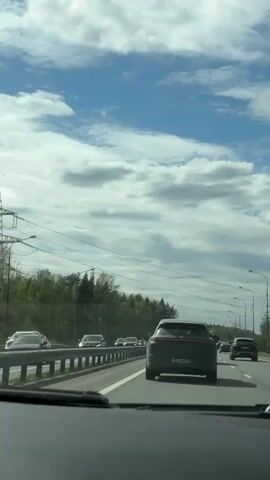 The video shows a view from inside a car traveling on a highway. The sky is blue with scattered clouds. Other cars are visible on the highway. A forest line is seen to the left. Power lines run parallel to the road.