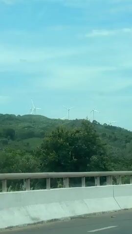 The video shows a view from a vehicle moving along a road. In the background, there is a green, hilly landscape with several wind turbines visible on the hilltops. Trees and vegetation cover the hills. In the foreground, a white barrier runs along the side of the road. A motorcycle with two occupants passes by. Further along the road, a lamppost stands beside the barrier, and a traffic sign with red and white markings is visible. The video also shows a green trash bin by the roadside and a building with a distinct roofline. The sky is blue with scattered clouds.