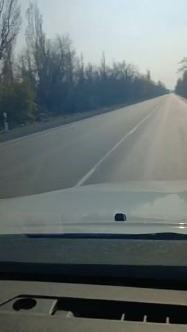 The video depicts a car driving down a two-lane road on a sunny day. The road is asphalt and has a single white line dividing the lanes. The sun is visible in the sky, causing a glare. Trees line both sides of the road, with bare branches suggesting it might be late fall or winter.  A box truck passes in the adjacent lane, moving in the same direction as the recording car. The dashboard of the car is visible in the foreground. The overall atmosphere is bright and clear.