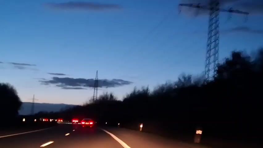 The video depicts a highway at dusk with several cars traveling in the same direction.  The sky is a gradient of blues, and a few clouds are visible.  Power lines and towers are visible along the side of the road, and the trees are silhouetted against the darkening sky.  The camera appears to be mounted inside a moving car.  The tail lights of the cars ahead are illuminated.  The white lane markings are visible on the road.  Music is playing in the background.