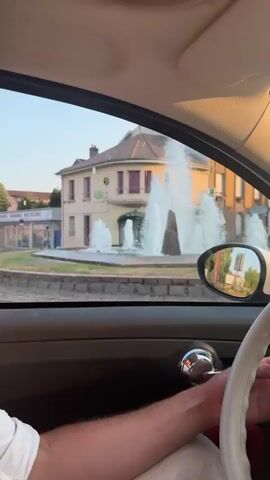 The video shows a point-of-view perspective from inside a car as it drives through a town. In the foreground, a person's arm is visible on the steering wheel and their shoulder. The interior of the car is visible, including the dashboard and part of the passenger door. Through the driver's side window, a large fountain with multiple water jets is visible in the center of a roundabout. Behind the fountain are buildings with shops and apartments. The side mirror reflects a similar scene of buildings and trees. As the car continues, the fountain recedes and the view shifts to a street lined with buildings and trees, with parked cars on the side of the road. The car passes a building with signage for "CIC" and what appears to be a town hall or municipal building. The street continues with more buildings and parked cars, and the sky is clear and appears to be late afternoon or early evening. The car is moving, indicated by the motion blur and the changing scenery.