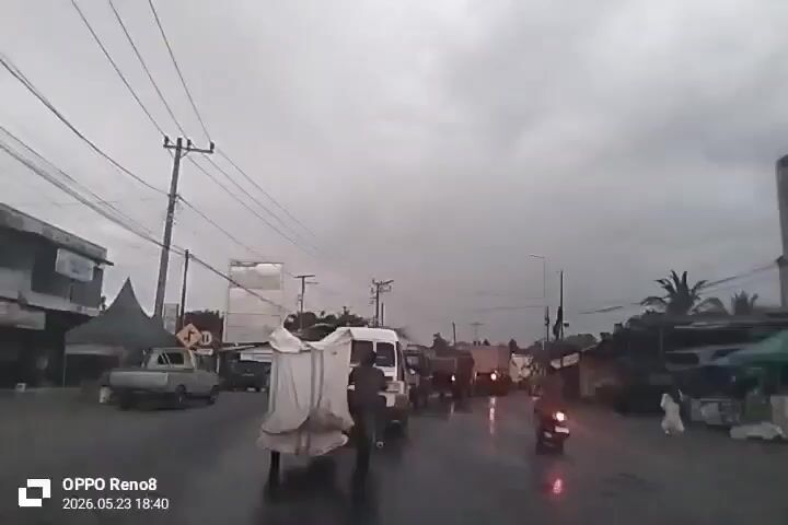The video shows a street scene on a rainy day. The sky is overcast and the road is wet. There are utility poles with power lines overhead. On the left side of the road, there is a parked pickup truck and a tent-like structure. A man is walking on the road, pushing a two-wheeled cart covered with a white tarp. A white van is driving behind him. A motorcycle is also on the road. On the right side of the road, there are buildings and some vegetation, including a palm tree. The bottom left corner of the screen has a timestamp and camera information.