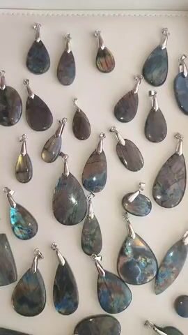 This is a close-up, vertical, overhead shot of a collection of labradorite pendants. The pendants are of various shapes and sizes, with a few appearing to be teardrop-shaped, while others are more elongated or oval. Each pendant is set with a silver bail at the top for attachment. The labradorite stones display their characteristic iridescent sheen, with flashes of blue, green, yellow, and purple visible against a darker, often gray or black, background. Some pendants exhibit more prominent color play than others. The pendants are arranged on a light-colored surface, possibly a display tray or a white fabric, with a slight shadow cast by some of the bail attachments. The lighting is bright, highlighting the natural beauty and unique optical properties of the labradorite. There is no visible text in the video. The overall impression is one of a collection of polished gemstones, likely for sale or display. The scene appears to be an indoor setting, possibly a jewelry store, market stall, or a home studio where these pendants are being showcased. The time of day and weather are not discernible from the provided imagery, and no people are visible.