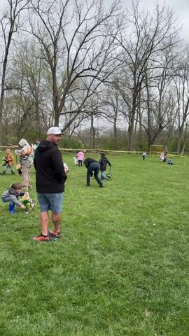 The video shows an Easter egg hunt taking place on a grassy field. Several children and adults are scattered across the field, looking for eggs. Some children are carrying baskets and picking up eggs, while adults are observing or helping. The background includes trees and a yellow caution tape marking the boundaries of the egg hunt area. The sky is overcast.