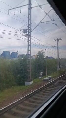 The video shows a view from inside a moving train. The train tracks are visible in the foreground, with the right side of the tracks blurred due to motion. To the left of the tracks, there is a green, vegetated area that gives way to a storage yard filled with shipping containers of various colors, including blue, white, and orange. Numerous utility poles and overhead wires run across the frame, suggesting the train is on an electrified line. The sky is overcast with clouds.