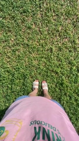 The video opens with a first-person perspective of someone standing on a green lawn. Their feet, clad in white sandals, are visible at the bottom of the frame. The person is wearing a pink t-shirt with text on it and a blue denim skirt. The camera then pans slightly to the right, showing more of the grassy area. The next scene is a wider shot of a brown stone church with a conical dome and a cross on top, set against a bright blue sky dotted with white clouds. The church is situated on a grassy knoll.