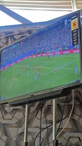 A television screen displays a soccer match. The game appears to be in progress, with players on the field and spectators in the stands. The screen shows a scoreboard or broadcast overlay with game information. The television is mounted on a stand. The background behind the television is a wall with a geometric pattern. The lighting suggests an indoor or shaded outdoor setting.