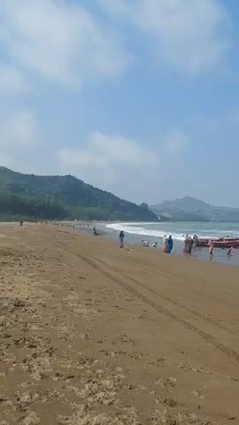 A wide shot of a sandy beach shows several people interacting near a red boat at the edge of the water. In the background, lush green hills rise against a bright blue sky with scattered clouds. The waves gently lap at the shore, creating white foam. Tire tracks are visible in the wet sand. A few individuals are scattered further down the beach, some appearing to be wading in the water. The overall scene depicts a leisurely day at the beach.