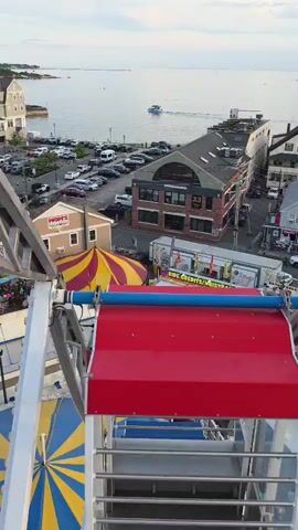This media file presents an aerial perspective, likely from a Ferris wheel or similar amusement ride, overlooking a bustling waterfront town during what appears to be a fair or carnival. The scene is set against a backdrop of the ocean and a cloudy sky, suggesting it is either early morning or late afternoon.

Prominently featured in the foreground is the structure of the ride itself, with its red and blue components. Below, the fairgrounds are teeming with people, indicating a lively event. Numerous tents, game booths, and food stalls are visible, contributing to the festive atmosphere. One stall has visible text that reads "RIDE CREDITS/WRISTBANDS". Cars are parked along the streets and in a designated lot, hinting at the number of attendees.

The town itself is a mix of buildings, including residential homes and commercial establishments, situated along the waterfront. A few boats are present in the water, with one motorboat actively moving. The overall impression is one of a vibrant community event taking place in a coastal setting. The lighting and cloud cover suggest a temperate climate, and the presence of a crowd implies good weather for an outdoor gathering. The perspective emphasizes the scale of the fair and its integration into the town's landscape.