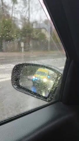 The video shows the perspective of a driver on a rainy day, with an ambulance moving in the opposite direction. The initial shot is through the side window, with raindrops obscuring the view of the yellow and red ambulance. As the video progresses, the view shifts to the windshield, showing the ambulance driving away in the distance, with its blue lights flashing. There is a blue car driving in the same direction as the driver, and trees line the sides of the road. The windshield wipers are active, clearing the rain. The road is wet and reflects the light.