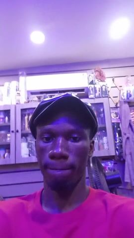 A close-up selfie video shows a young, dark-skinned man wearing a red shirt and a black cap, looking directly at the camera with a neutral expression. The background appears to be a retail store or salon, with shelves filled with various products visible behind him. The lighting in the room is primarily purple, creating an overall violet tint.