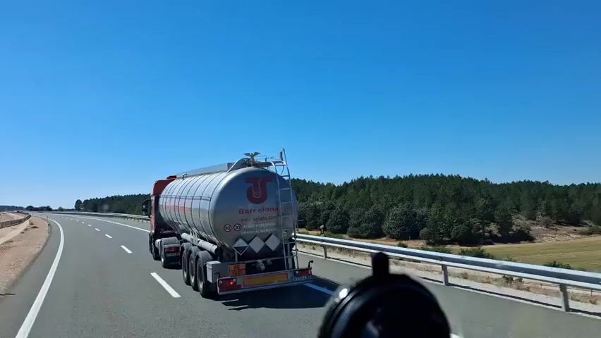 A tanker truck drives down a highway under a clear blue sky. The truck has a silver tank with a red logo that says "Barcelona" and "solazo.com". It is also marked with speed limits of 80 and 90 and shows hazard symbols. The truck is surrounded by trees and fields. The camera is positioned inside a vehicle, looking out at the tanker truck.