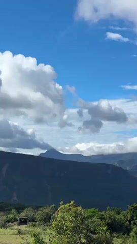 The video shows a mountain range covered in green vegetation, with a cloudy blue sky overhead. The clouds are white and fluffy, and they partially obscure the peaks of the mountains. The camera pans slowly across the landscape, providing a wide view of the scene. There are no people or animals visible in the video. The environment appears to be a natural one, with no signs of human development.