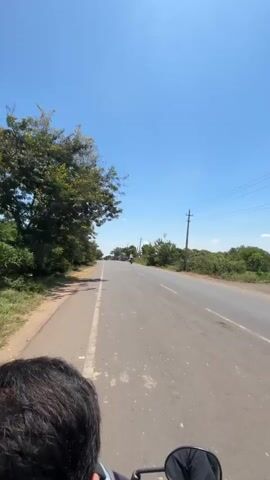 The video begins from the perspective of someone riding a scooter, looking forward along a paved road. A person's dark hair is visible in the foreground. The road is bordered by trees and vegetation on the left, and more sparse vegetation, a utility pole, and some buildings in the distance on the right. The sky is clear and blue. As the video progresses, a white SUV appears on the right side of the road, also traveling in the same direction. Further down the road, several motorcycles are visible. The overall environment appears to be a rural or semi-rural area on a sunny day.