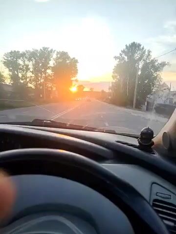 The video shows a first-person view from inside a car looking out the windshield at a sunset. The sun is a bright orange orb low on the horizon, casting a warm glow over the scene. Trees line the sides of the road, and power lines are visible in the distance. There are several cars on the road ahead, moving away from the viewer. A hand is visible gripping the steering wheel, indicating that the person is driving. The sky is a pale blue with some scattered clouds. The overall atmosphere is peaceful and serene.