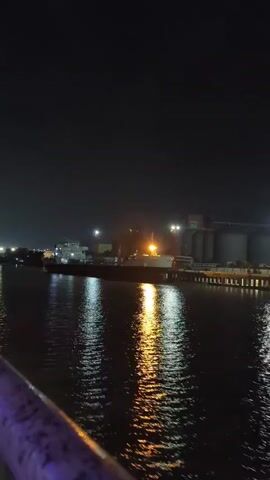 A video captures a night scene at a port. A large cargo ship is docked, illuminated by lights. The water in front of the ship reflects the lights, creating a shimmering effect. The sky is dark, indicating nighttime. In the background, there are port facilities and other structures. The camera appears to be moving slightly, suggesting it is mounted on a vehicle or being held by someone. There are no people visible in the video.