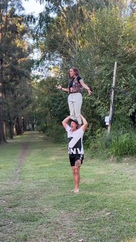 A woman stands on a man's shoulders in a grassy park, both facing forward and smiling. The man wears a hat, a black and white t-shirt with lettering, black shorts, and is barefoot. The woman wears a black crop top, patterned vest, and beige pants. A narrow dirt path leads into a forested area behind them. A dog sits in the lower left corner. The camera pans up to show a drone tangled in the branches of a tall tree. High-voltage power lines are visible above the tree branches.