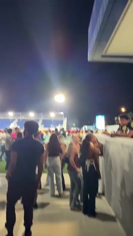 The video shows a crowd of people standing outside of a stadium at night. The stadium is lit up with bright lights, and there is a large screen displaying images on the outside of the stadium, visible behind the people. There is a police officer standing at a counter or window on the right side of the frame, talking to a few of the people in the crowd. The people are dressed in casual clothing, and they appear to be waiting to enter the stadium or gathering before or after an event. Music is playing in the background. The environment is lively with the crowd and the lights of the stadium.