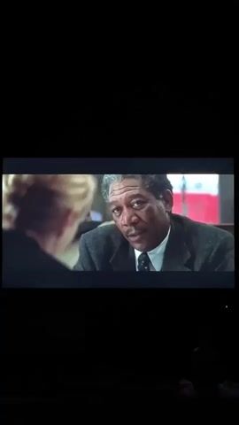 The scene shows Morgan Freeman in a suit speaking with a woman. The woman sits with her hands folded in front of her. They are seated in a booth inside what looks like a diner. Other people are seated in the background.