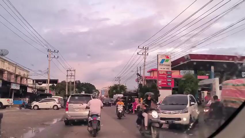 The video shows a street scene in front of a Pertamina gas station. There are several cars and motorcycles on the road. The sky is overcast. Power lines run across the street. The gas station is red and white. There are people on motorcycles and in cars. Some people are wearing helmets. The road is wet.
