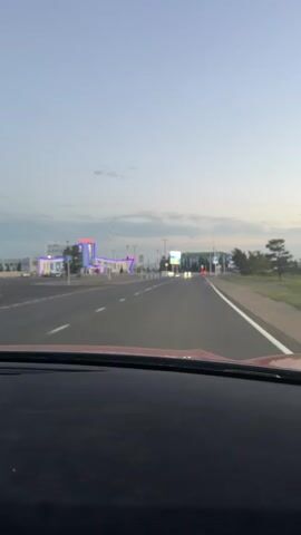 The video shows a view from inside a vehicle traveling on a road. The road has white lane markings and is flanked by a sidewalk and fences. On the left side of the road, there is a modern-looking building with blue and white accents and neon lights. The building appears to be a public facility, possibly a train station or bus terminal, as suggested by the signage with Cyrillic lettering. There are streetlights and trees along the road and in front of the building. In the distance, other buildings and lights are visible. The sky is overcast and appears to be either dawn or dusk. There are no people clearly visible in the video. The vehicle is moving forward, and the camera captures the scene as it passes by.
