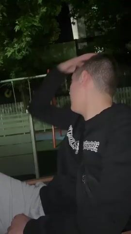 A young man sits on a wooden bench at night. He is wearing a black long-sleeved shirt with white text and grey sweatpants. His left hand is partially covering his mouth and nose, and he is looking to the right side of the frame. In the background, there are green trees, a dark building, and some playground equipment visible in the low light.
