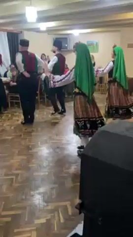 In a medium shot indoors, two men and two women are shown performing a traditional folk dance. The men are wearing black caps, black pants, white long-sleeved shirts, and vests with a red and green design. The women are in traditional dresses, with green headscarves, embroidered tops, and plaid skirts with floral patterns. The dance involves coordinated steps and arm movements. The room has a wooden floor, and there are tables and chairs with people sitting at them in the background, observing the performance. A large black speaker is in the foreground on the right. There are ceiling lights and paintings on the walls.