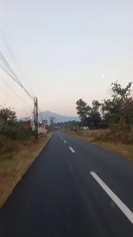 This is a first-person point-of-view video taken from a vehicle traveling down a paved road. The road has white dashed lines marking the lanes. On the left side of the road, there are power lines, a utility pole, some buildings, and dry vegetation. On the right side of the road, there are trees and some houses or buildings in the distance. In the background, there is a mountain or large hill under a pale sky. The time of day appears to be either sunrise or sunset.