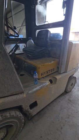 The video shows a static, close-up shot of a yellow Hyster forklift parked inside what appears to be a garage or warehouse. The forklift is dirty and shows signs of wear. A blue water bottle sits on its side near the "HYSTER" logo. The seat is visible, and the window behind the seat provides a view of an outdoor area with a few buildings and a muddy ground. The sky is visible in the background, appearing blue. The camera doesn't move throughout the video.