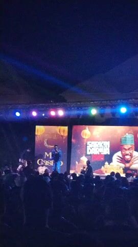 The scene captures a live stage performance in front of a crowd. On the stage, a man dressed in jeans and a jacket is visible, holding a microphone and actively performing. Two large screens behind him display graphics, one with the words "Mc Casino" and the other featuring a portrait of a man wearing a traditional hat with "Sarkin Dariya" written on it.  Several other individuals are positioned around the main performer on stage. The audience is visible in the foreground, with their silhouettes showing they are closely observing the performance. The lighting consists of several spotlights above the stage, creating a mix of blue and white illumination.  The overall atmosphere suggests a comedy show or a festive event.